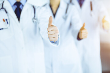 Wall Mural - Group of unknown doctors stand as a team with thumbs up in a sunny hospital office. Physicians ready to examine and help patients. Medical help, insurance in health care, best desease treatment and me