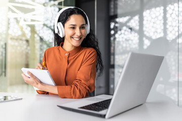 latin american business woman with curly hair and headphones watching online training course at work
