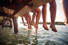 Friends Sitting On The Dock And  Enjoying Together At Vacation Dangling Legs In The Water