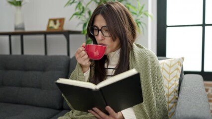 Poster - Young beautiful hispanic woman reading book and drinking coffee sitting on sofa at home