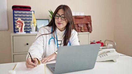 Wall Mural - Young beautiful hispanic woman doctor having video call writing notes at clinic