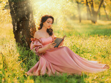 Fantasy Woman Sits Under Tree Holding Romantic Book In Hands Reading Novel. Pink Long Vintage Dress. Fairy Princess Girl In Garden Summer Nature Green Grass Magic Sun Rays Light. Art Photo Real People