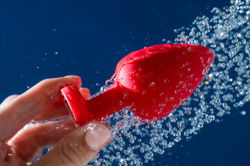 Woman washing red anal plug under shower on blue background. 