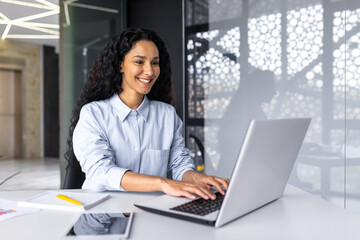 Wall Mural - Happy and smiling hispanic businesswoman typing on laptop, office worker with curly hair and glasses happy with achievement results, at work inside office building.