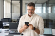 © Stock 4 You - Close-up of smiling mature Latin or Indian businessman holding smartphone in office. Middle aged manager using cell phone mobile app. Digital technology application and solutions for business concept.