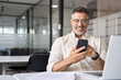 © Stock 4 You - Middle aged Hispanic business manager ceo using cell phone mobile app, laptop. Smiling Latin or Indian mature man businessman holding smartphone sit in office working online on gadget with copy space.