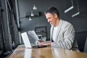 Middle aged happy professional business man company executive manager wearing suit sitting at desk in office working checking bills corporate financial accounting documents using laptop computer.