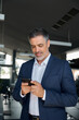 © insta_photos - Happy mid aged business man ceo wearing blue suit standing in office using cell phone. Mature businessman professional executive manager holding mobile checking applications on cellphone. Vertical