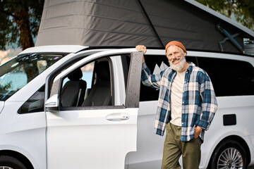 Wall Mural - Happy older man standing with rv camper van on travel vacation. Smiling mature active traveler advertising campervan car with tent on roof enjoying camping tourism road trip, campervan booking rental.