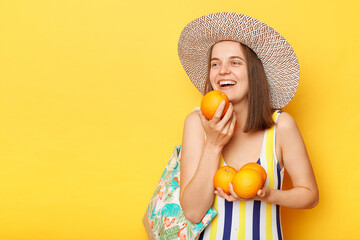 Dreamy happy joyful woman wearing swimsuit isolated on yellow background holding oranges looking away with optimistic expression empty space for promotional text.