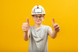 Guy with glasses wears protective helmet and shows thumbs up with two hands. Teenager shows gesture like isolated on yellow background.