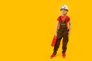 Wall Mural - Boy imitates worker or construction worker. Child in work overalls and helmet with suitcase of tools. Mock up, copy space