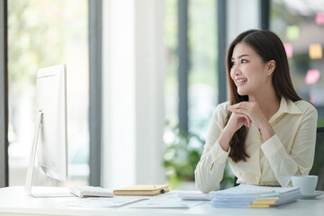 Photo of attractive woman sitting at desk working on computer looking at screen, smiling, wearing yellow shirt and glasses, typing on keyboard, holding a cup of coffee.