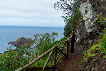 Wall Mural - Young blonde woman with her back on the turquoise ocean waves, cliffs and rocks background. Travel girl photography. Beautiful landscape. Madeira island, Portugal.