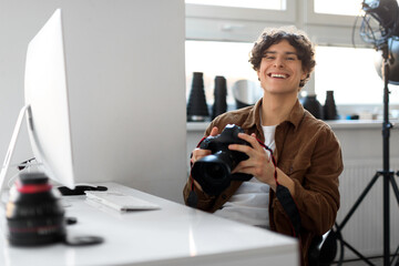 Wall Mural - Professional photographer young man working on photo shoot at studio, holding dslr camera, using pc computer