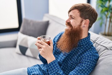 Poster - Young redhead man drinking tea sitting on sofa at home