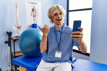 Poster - Young man working at rehabilitation clinic doing video call with smartphone pointing thumb up to the side smiling happy with open mouth