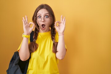 Young caucasian woman wearing student backpack over yellow background looking surprised and shocked doing ok approval symbol with fingers. crazy expression