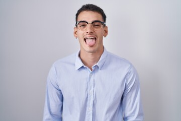 Wall Mural - Handsome hispanic man wearing business clothes and glasses sticking tongue out happy with funny expression. emotion concept.