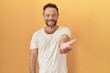 © Krakenimages.com - Middle age man with beard standing over yellow background smiling cheerful offering palm hand giving assistance and acceptance.