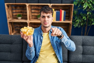 Sticker - Young hispanic man holding potato chips pointing with finger to the camera and to you, confident gesture looking serious