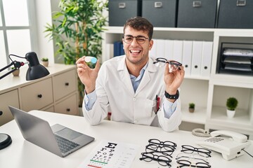 Sticker - Young hispanic oculist holding glasses and contact lenses winking looking at the camera with sexy expression, cheerful and happy face.