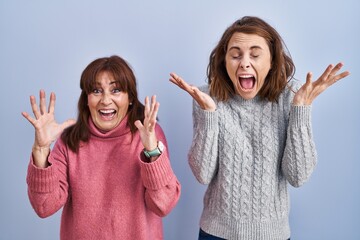 Poster - Mother and daughter standing over blue background celebrating mad and crazy for success with arms raised and closed eyes screaming excited. winner concept