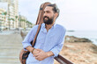 Young hispanic man musician holding guitar case leaning on balustrade at seaside