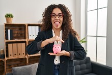 Young Hispanic Woman Holding Piggy Bank Sticking Tongue Out Happy With Funny Expression.