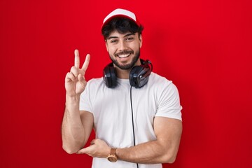 Wall Mural - Hispanic man with beard wearing gamer hat and headphones smiling with happy face winking at the camera doing victory sign with fingers. number two.