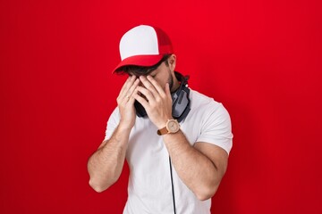 Sticker - Hispanic man with beard wearing gamer hat and headphones with sad expression covering face with hands while crying. depression concept.