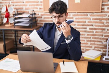 Wall Mural - Young hispanic man business worker using laptop talking on smartphone at office