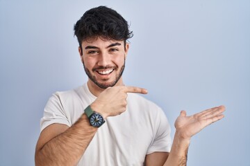 Canvas Print - Hispanic man with beard standing over white background amazed and smiling to the camera while presenting with hand and pointing with finger.