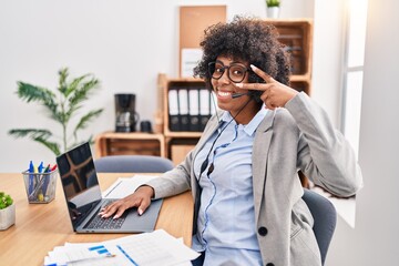 Canvas Print - Black woman with curly hair wearing call center agent headset at the office doing peace symbol with fingers over face, smiling cheerful showing victory