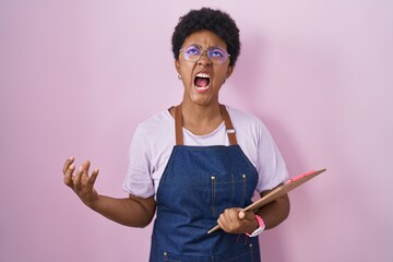 Wall Mural - Young african american woman wearing professional waitress apron holding clipboard angry and mad screaming frustrated and furious, shouting with anger. rage and aggressive concept.