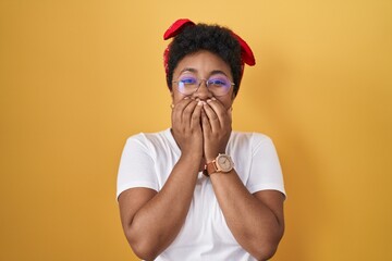 Poster - Young african american woman standing over yellow background laughing and embarrassed giggle covering mouth with hands, gossip and scandal concept