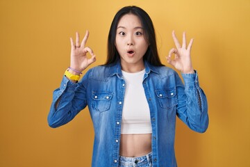 Poster - Young asian woman standing over yellow background looking surprised and shocked doing ok approval symbol with fingers. crazy expression