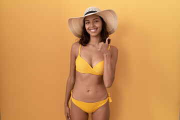 Wall Mural - Young hispanic woman wearing bikini and summer hat doing happy thumbs up gesture with hand. approving expression looking at the camera showing success.