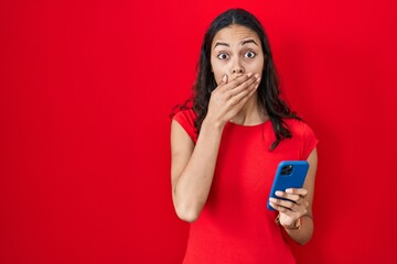 Canvas Print - Young brazilian woman using smartphone over red background shocked covering mouth with hands for mistake. secret concept.