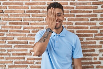 Wall Mural - Brazilian young man standing over brick wall covering one eye with hand, confident smile on face and surprise emotion.