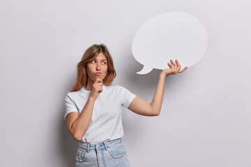 Pensive young European woman holds chin and looks doubtful holds blank speech communication bubble thinks what text to write dressed in casual t shirt and jeans isolated over white background.