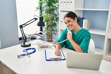 Canvas Print - Young beautiful hispanic woman doctor talking on telephone writing on document at clinic