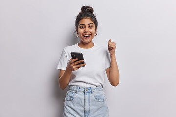Happy cheerful Indian girl with dark hair combed in bun clenches fist celebrates success holds smartphone dressed in casual basic t shirt jeans isolated over white wall receives message about reward