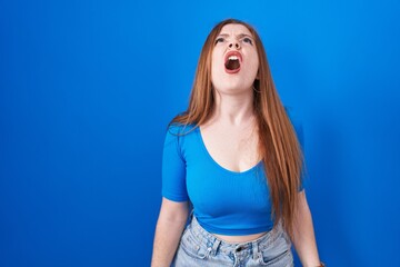 Canvas Print - Redhead woman standing over blue background angry and mad screaming frustrated and furious, shouting with anger. rage and aggressive concept.