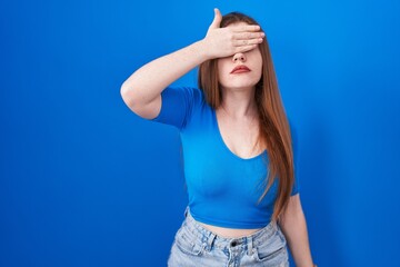 Wall Mural - Redhead woman standing over blue background covering eyes with hand, looking serious and sad. sightless, hiding and rejection concept