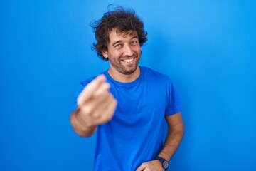 Poster - Hispanic young man standing over blue background beckoning come here gesture with hand inviting welcoming happy and smiling