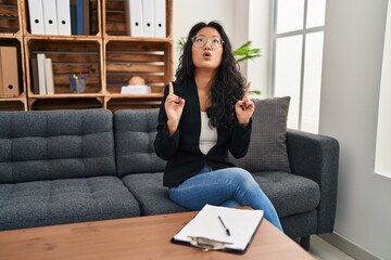 Canvas Print - Young asian woman at consultation office amazed and surprised looking up and pointing with fingers and raised arms.