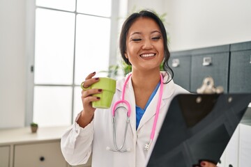 Wall Mural - Young chinese woman wearing doctor uniform holding clipboard drinking coffee at clinic