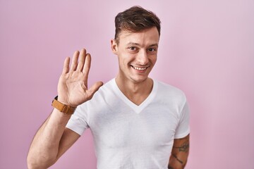 Caucasian man standing over pink background waiving saying hello happy and smiling, friendly welcome gesture