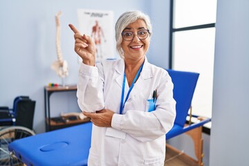 Canvas Print - Middle age woman with grey hair working at pain recovery clinic with a big smile on face, pointing with hand finger to the side looking at the camera.